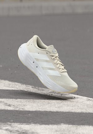 Zapatilla deportiva beige claro con rayas blancas de Adidas, flotando sobre una carretera de asfalto gris con marcas blancas de carril y pequeños escombros negros.