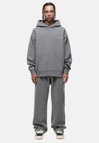Persoon met gevlochten haar, gekleed in een grijze oversized hoodie en bijpassende joggingbroek, staand tegen een eenvoudige witte achtergrond.
