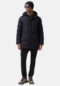 Mand iført en sort puferjakke med hætte, sort beanie, solbriller, mørke bukser med sidelommer og brune og grå sneakers, som står med front mod kameraet.
