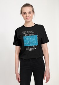 Usmívající se žena v černém tričku s motivem Picassa, na kterém je modrá grafika s abstraktními tanečními postavami a bílý text, v neformálním studiu.