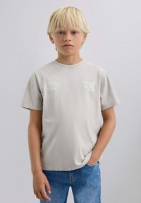 T-shirt en coton gris clair à manches courtes, arborant un motif de globe et une impression de texte sur la zone de la poitrine gauche. Associé à un jean en denim bleu.