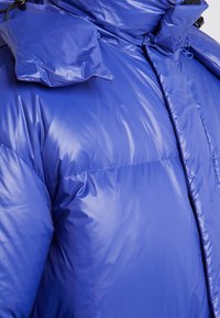 Jasně modrá péřová bunda s lesklým povrchem, s vysokým límcem a odnímatelnými ramenními vycpávkami. Zapínání na zip a šité designové detaily.