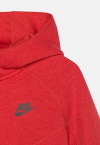Czerwona bluza Nike z kapturem, wykonana z teksturowanej tkaniny, z wysokim kołnierzem i małym czarnym logo z przodu. Projekt zawiera kontrastowe szwy.