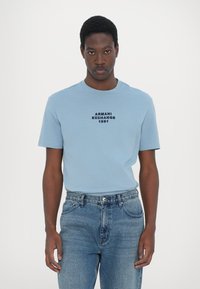 Svijetlo plava majica s kratkim rukavima i okruglim izrezom, s tamnoplavo otisnutim logotipom "ARMANI EXCHANGE 1991", u kombinaciji s plavim trapericama.
