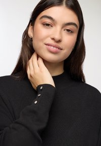 Maglione nero a coste con collo alto e bottoni decorativi sulle maniche. Texture morbida e vestibilità rilassata in evidenza.