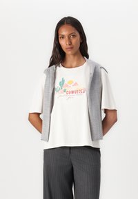 Short-sleeve white t-shirt with a graphic print featuring cacti and mountains, layered under a gray sleeveless vest. Gray pinstripe trousers.