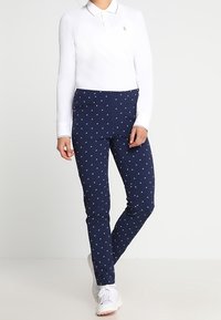 Vit långärmad polo tröja i kombination med marinblå byxor med ett litet upprepat mönster. Outfitten inkluderar ljusa sneakers.