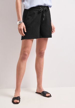 Femme portant un short noir à cordon, un haut bleu clair, des sandales slides noires et des bracelets, debout devant un fond clair uni.