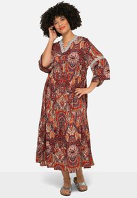 Vestido maxi fluido con estampado de paisley multicolor. Presenta mangas acampanadas, un escote con forma de ola y un tejido ligero. Sandalias plateadas visibles.