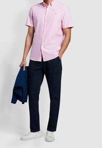 Camicia rosa a maniche corte con bottoni, pantaloni blu scuro e sneakers bianche. La camicia presenta un logo sottile e un design del colletto classico.
