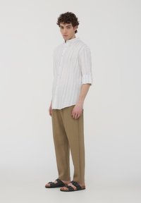 Camicia bianca a righe con bottoni, pantaloni beige a fit morbido, sandali neri con cinturini regolabili. Texture del tessuto liscia, taglio moderno.