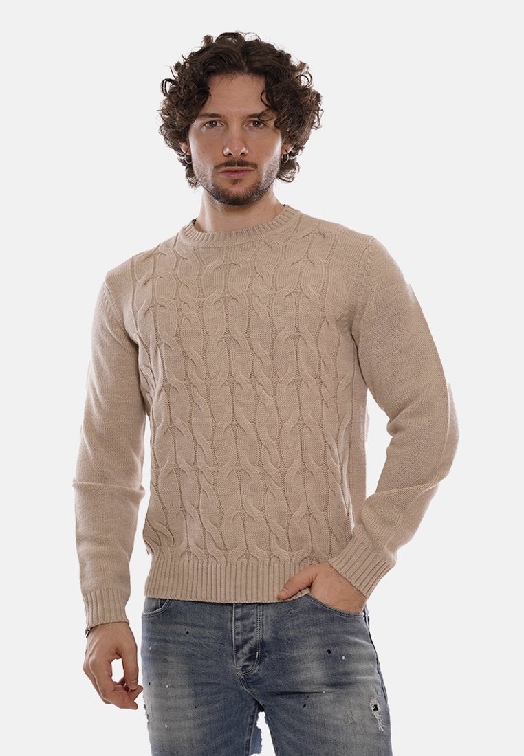 Uomo con capelli ricci che indossa un maglione beige a trecce e jeans blu strappati, in piedi con una mano in tasca contro uno sfondo bianco.