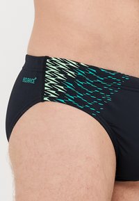 Svart simbrief med ett dynamiskt grönt mönster på sidan. Har en tajt passform och en slät yta. Logotyp på baksidan.