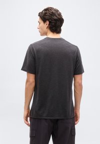 Νεαρός με κ οντά σγουρά μαλλιά που φορεί σκούρο γκρι κοντομάνικο μπλουζάκι και μαύρο παντελόνι cargo, στέκεται με την πλάτη προς τα εμπρός σε απλό φόντο.