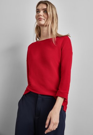 Top de manga larga rojo con relieve, con un escote amplio y puños enrollados, combinado con pantalones azul marino. Textura suave, diseño casual.