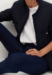 Námořnický modrý strukturovaný blazer s třepícími se okraji, s stříbrnými knoflíkovými akcenty, nošený přes bílý tričko a tmavě modré džíny. Póza vsedě.