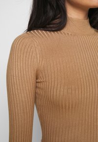 Tan ribbet strikket topp med høy hals og lange ermer, med en teksturert overflate og en tettsittende silhuett.