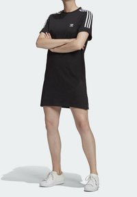 Personne portant une robe T-shirt noire Adidas avec des bandes blanches sur les épaules et des baskets blanches, se tenant les bras croisés.