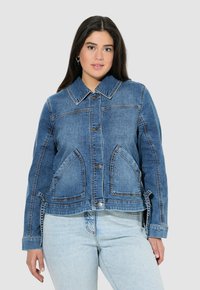 Džínová bunda ve středně modré barvě s límečkem, zapínáním na knoflíky a předními kapsami. Má kontrastní švy a boční šňůrky.