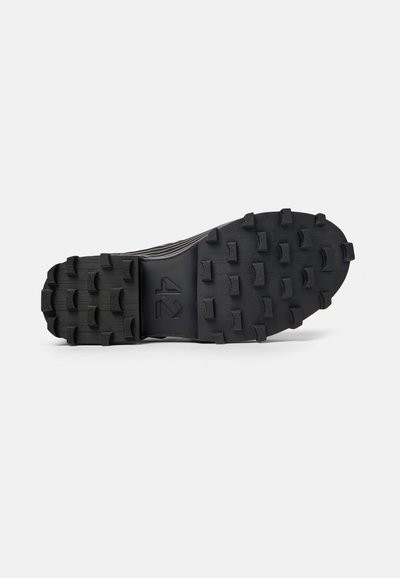 Semelle extérieure de chaussure de sport noire présentant une surface texturée avec des crampons profonds et agressifs pour une meilleure traction. Talon plat avec un détail de marque central.