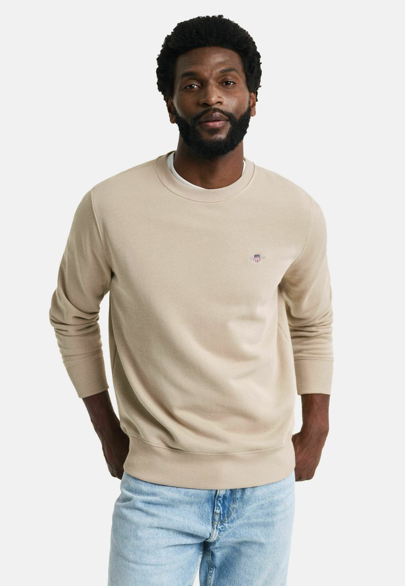 Beige sweatshirt met geribbelde ronde hals en manchetten, voorzien van een subtiele logo op de borst. Gecombineerd met lichtblauwe spijkerbroek.