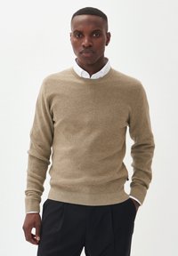 Suéter de punto beige con cuello redondo y puños acanalados, llevado sobre una camisa blanca con cuello. Unos pantalones negros completan el conjunto.