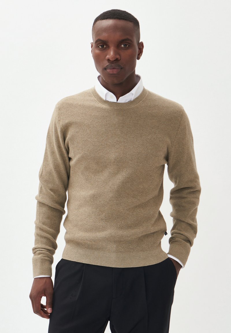 Suéter de punto beige con cuello redondo y puños acanalados, llevado sobre una camisa blanca con cuello. Unos pantalones negros completan el conjunto.