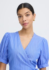 Femme aux cheveux foncés attachés en un chignon bas, portant des boucles d'oreilles créoles dorées et un chemisier bleu à manches courtes, col en V, à rayures, avec des épaules bouffantes.