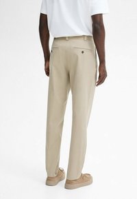 Uomo in pantaloni beige a vita alta, camicia bianca a maniche corte e scarpe in suede color sabbia in piedi su uno sfondo bianco.