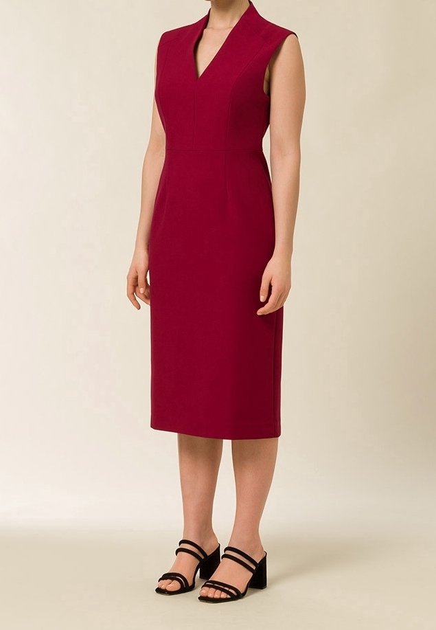 Femme portant une robe rouge foncé sans manches, longueur genou, avec un col en V et des sandales noires à talons blocs et lanières, debout devant un fond uni.