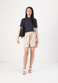 Top bleu marin cu dungi, cu mânecă scurtă, pantaloni scurți bej cu brâu și sandale albe. Modelul ține o geantă neagră, stând pe un fundal simplu.