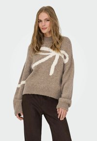 Pull en tricot marron avec un col montant et un grand motif abstrait blanc. Il est doté de poignets et d'un ourlet côtelés, avec une coupe ample. Rehaussé d'une texture douce.