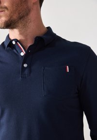 Marineblå pikéskjorte med en to-knapp placket, hvite knapper og en brystlomme med røde, hvite og blå detaljer. Strukturlignende stoff.