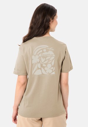 Femme aux longs cheveux bruns portant un t-shirt beige avec un motif floral et naturel et le mot « ensemble » au dos.