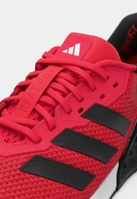 Red athletic shoe featuring a mesh upper, black accents, and textured laces. Includes a branded tongue label and cushioning sole design.