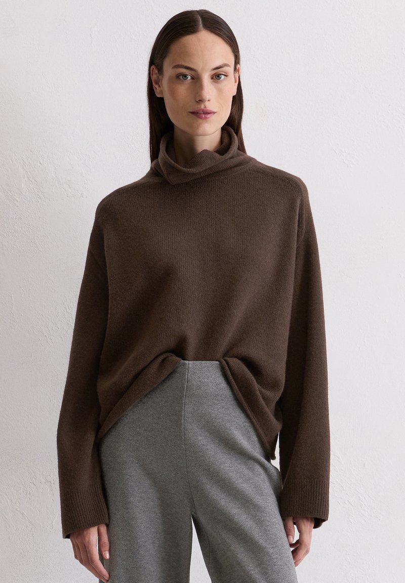 Brązowy sweter dzianinowy z wysokim kołnierzem, szerokimi rękawami i luźnym krojem, w zestawieniu z szarymi spodniami o wysokim stanie i zwężanym kroju.