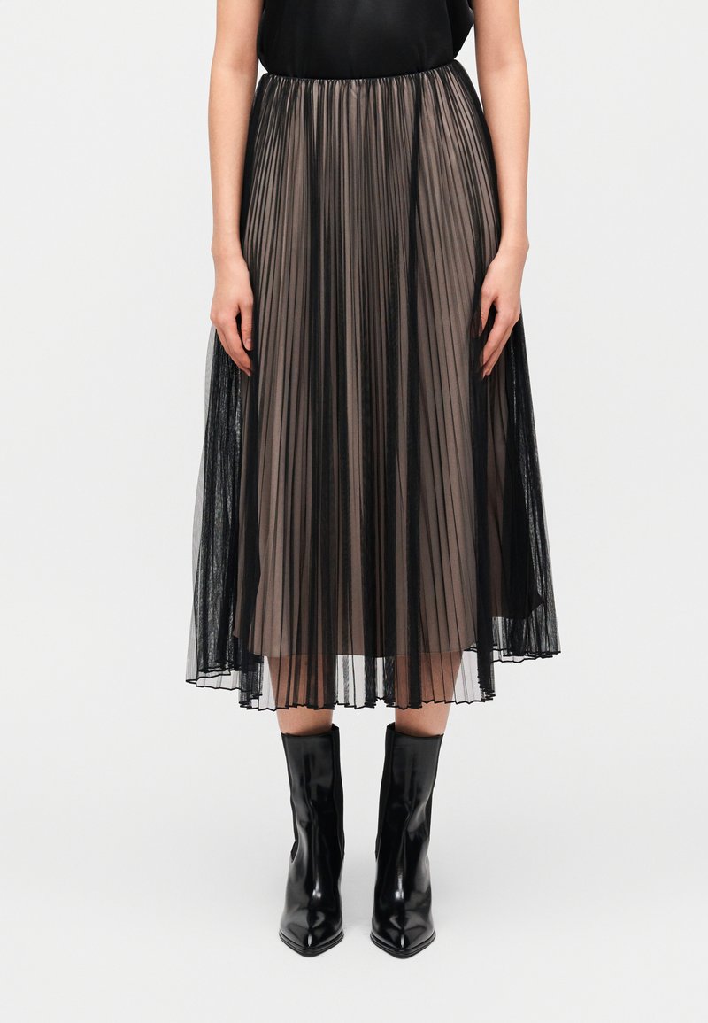 Πλισέ midi φούστα από διάφανο μαύρο και γκρι τούλι, με ελαστική μέση και αέρινη σιλουέτα, συνδυασμένη με μαύρα μποτάκια με μυτερή μύτη.