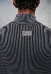 Een donker gestreepte sweater met een hoge kraag en een gestructureerde stof. Een klein label aan de achterkant toont "Vitoria" in een decoratief lettertype.