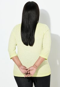 Jasnożółta, teksturowana koszula z rękawami do łokci, noszona z ciemnymi dżinsami, widziana od tyłu. Widoczne krótkie, proste czarne włosy.