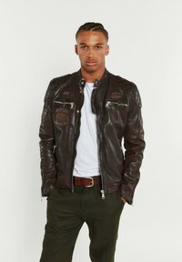 Veste aviateur en cuir marron avec des fermetures éclair, des accents épaules matelassés et des patchs brodés. Présente une coupe ajustée et une texture lisse.