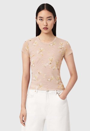 Ung kvinde med glat mørkt hår iført en gennemsigtig ferskenfarvet top med korte ærmer og blomsterprint samt hvide højtaljede bukser med den ene hånd i lommen.