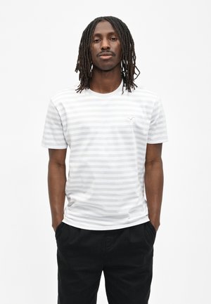 Mann mit Dreadlocks, der ein weißes und hellgrau gestreiftes Kurzarmhemd und schwarze Hosen trägt, mit den Händen in den Taschen vor weißem Hintergrund.