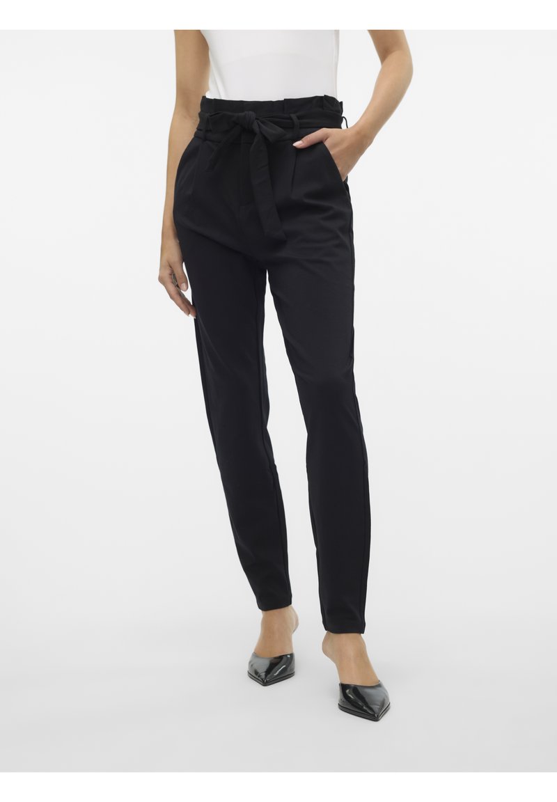 Femme portant un pantalon noir taille haute avec une ceinture nouée, associée à des escarpins noirs à bout pointu, debout sur un fond blanc.