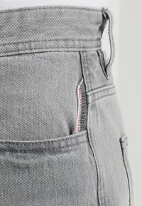 Jean en denim gris à taille haute, doté d'une poche latérale avec une couture rouge contrastante et d'une boucle à l'arrière. Texture lisse.