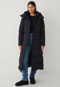 Lang sort dunjakke med hætte, der har et quiltet design, knaplukninger og sidelommer. Båret over en sort top og jeans.