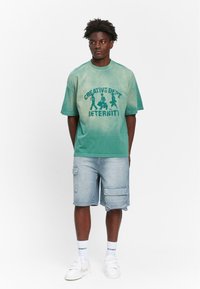 Grønfarvet oversized t-shirt med "CREATIVE DEPT ETERNITY" tryk, parret med lyseblå cargo shorts, hvide sneakers og sokker.