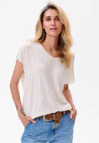 Weißes Kurzarmoberteil mit einem V-Ausschnitt, das ein strukturiertes Blumendesign aufweist. Kombiniert mit blauen Jeans und einem braunen Gürtel mit silbernen Akzenten.