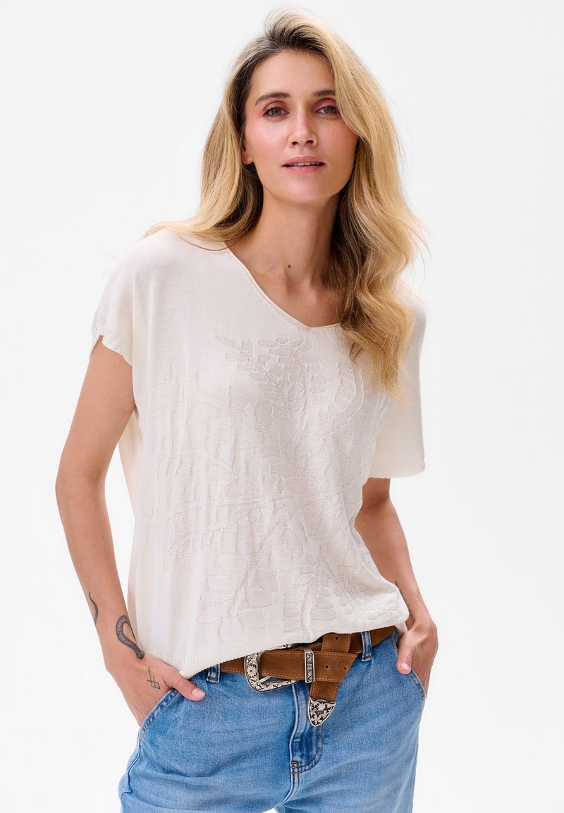 Weißes Kurzarmoberteil mit einem V-Ausschnitt, das ein strukturiertes Blumendesign aufweist. Kombiniert mit blauen Jeans und einem braunen Gürtel mit silbernen Akzenten.