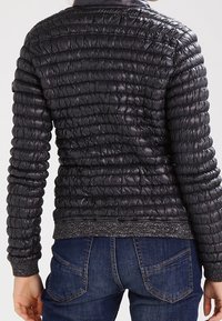 Zwarte pufferjack met een gequilt ontwerp, geribde manchetten, een elegante kraag en een metallic-accentrand, gecombineerd met blauwe jeans.