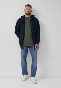 Marineblauwe zip-up hoodie met een voork pocket, gecombineerd met een olijfgroene t-shirt en lichtblauwe jeans, afgewerkt met witte sneakers.